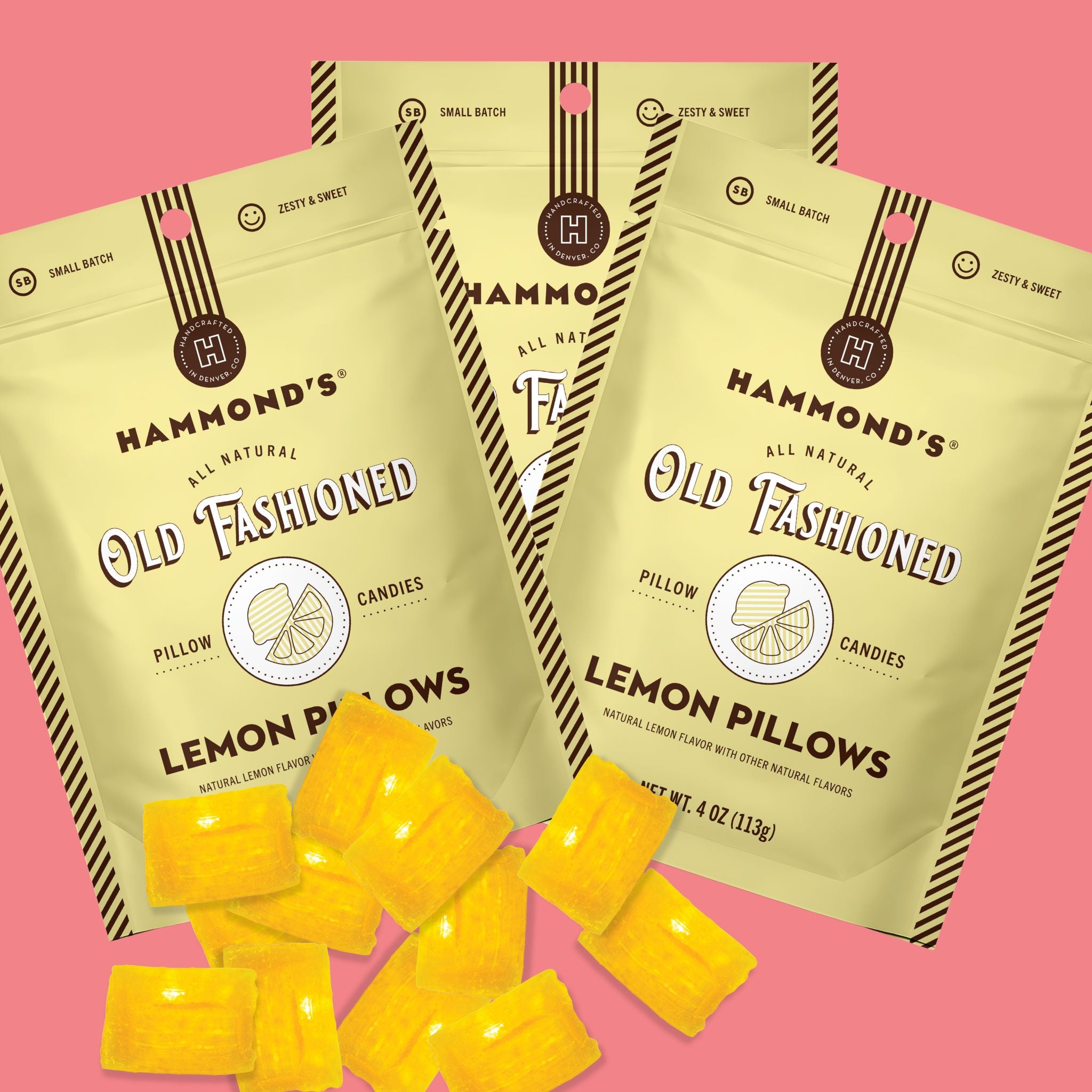 Three bags of Hammond's Old Fashioned Lemon Pillows candies are arranged on a pink background with several yellow gummy candies scattered in front. The packaging is pale yellow with dark brown text and striped accents, and the candies are bright yellow.