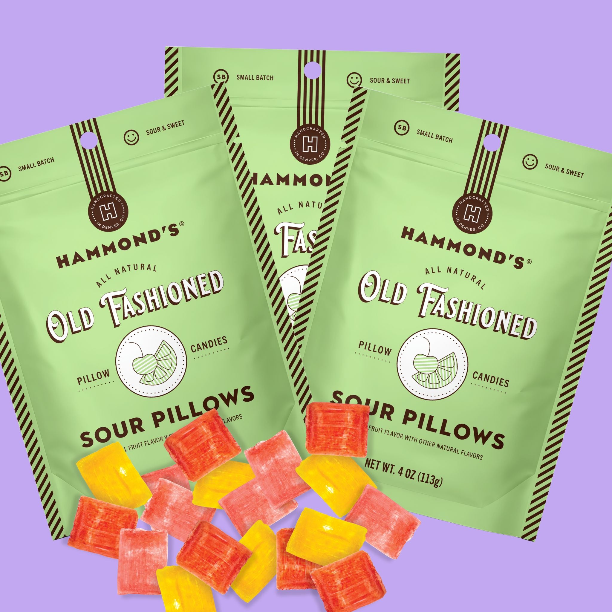 Three bags of Hammond's Old Fashioned Sour Pillows candy are arranged on a light purple background, with several red and yellow candy pieces scattered in front. The bags are light green with dark green and brown striped details, and the brand name Hammond's is clearly visible.