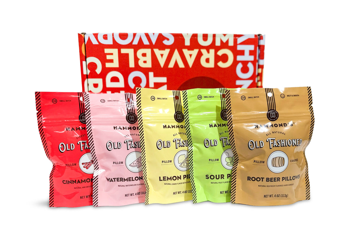 Four stand-up pouches of Old Fashioned Cravable Pillow faces are arranged in front of a red gift box on a white background. The pouches are red, pink, yellow, and tan, representing Cinnamon, Watermelon, Lemon Pie, and Sour Pie flavors.