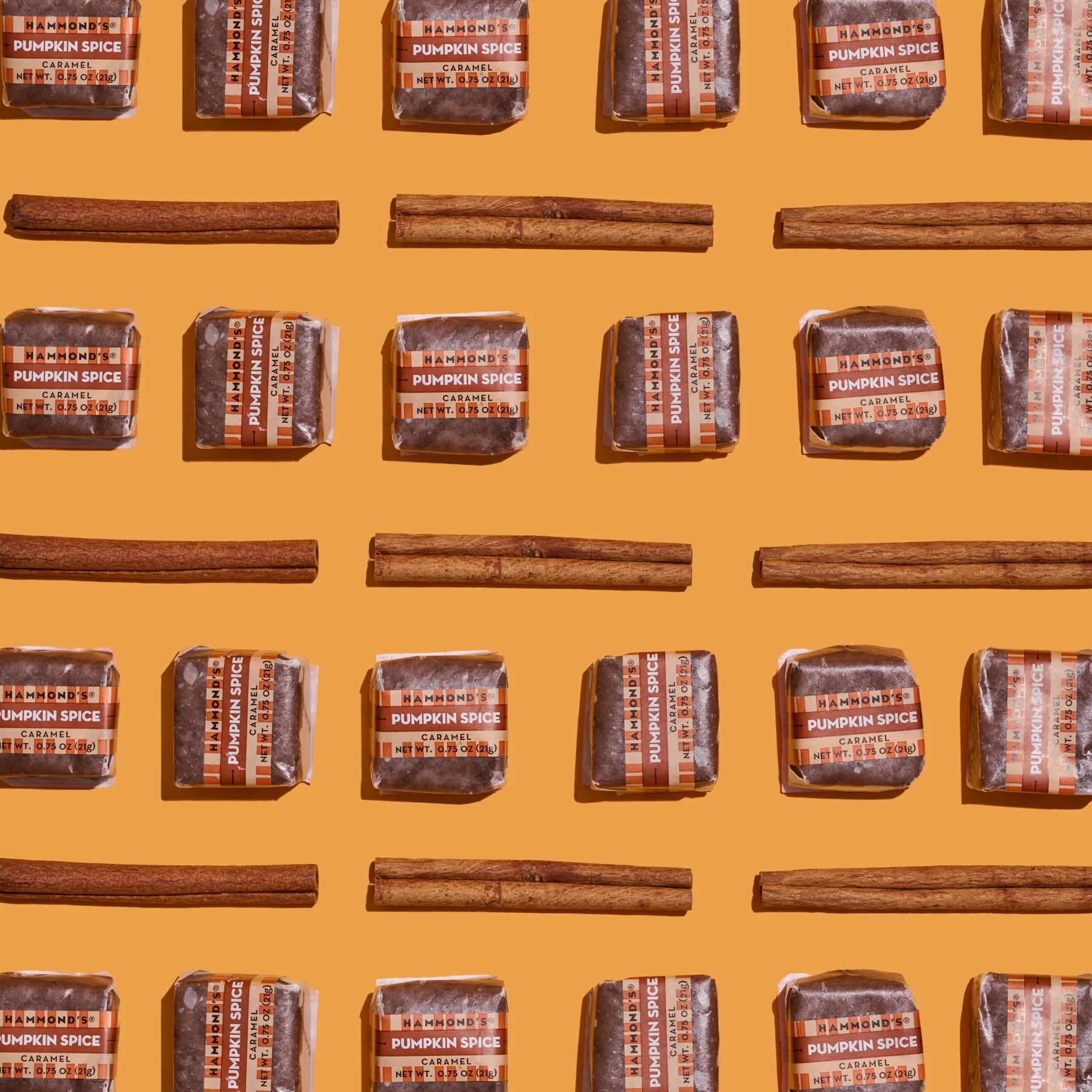 Individual pumpkin spice caramels laid flat in line with lines of cinnamon sticks on orange background